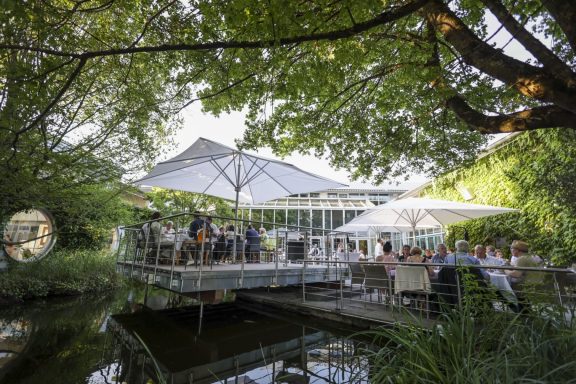Gemütliches Restaurant im Freien, umgeben von Bäumen und einem Wasserlauf.